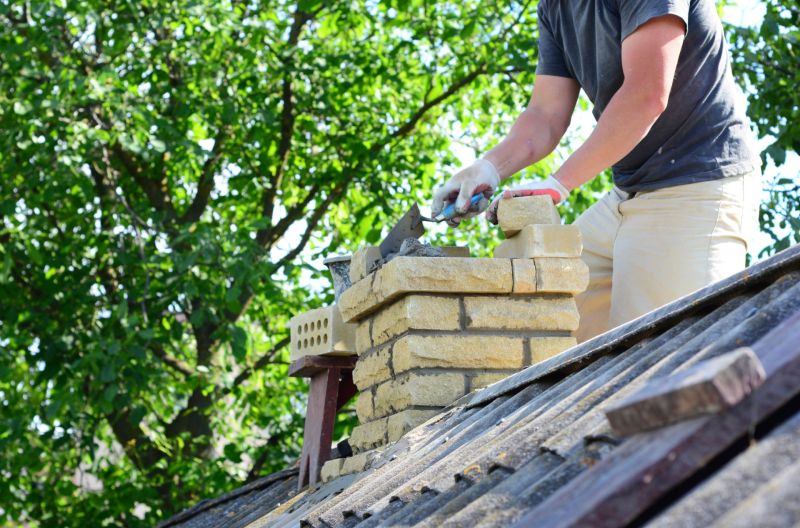 Worker repairing brick chimney on a sloped roof with trowel and mortar